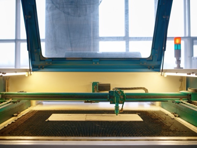 Grabadora láser con la tapa abierta: el pórtico y el cabezal cruzando la cama de panal, con una plancha lista para cortar y las luces del interior encendidas.